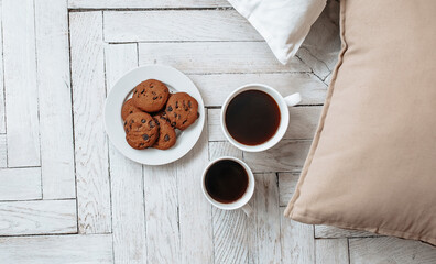 Breakfast in the morning. Two white cups of coffee and chocolate chip cookies stand on a light wooden parquet and pillows lie nearby. Homeliness, day off. Pastel colors.