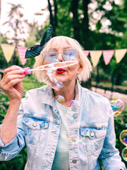 senior (old) stylish woman with gray hair and in blue glasses and jeans jacket blowing bubbles outdoors. Holidays, party, anti age, fun concept