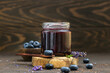 Glass jar and spoon with delicious blueberry jam with fresh berries, lavender and rosemary on dark brown table. Healthy food for breakfast.