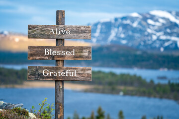 alive blessed grateful text on wooden signpost outdoors in landscape scenery during blue hour. Sunset light, lake and snow capped mountains in the back.