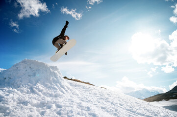 Stylish young girl snowboarder does the trick in jumping from a snow kicker against the blue sky clouds and mountains in the spring.