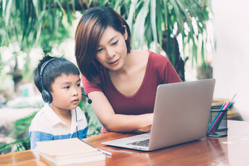 Wall Mural - Young asian mother and son using laptop computer for study and learning together at home, boy wearing headphone for e-learning with distancing, teacher or mom support child, education concept.