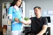 © H_Ko - Doctor feel hand in hospital. Man sit on chair with his arm extended forward with dumbbell at doctor appointment. Woman in doctor suit and red stethoscope examine visitor hand for fracture