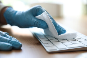 Woman in latex gloves cleaning computer keyboard with wet wipe at table, closeup