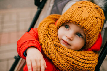 Wall Mural - Little cute baby with brown knitted hat and scarf lies in the stoller and looks aside