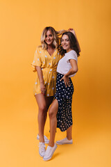 Amazing blonde girl in yellow outfit chilling with best friend. Indoor full-length shot of two enthusiastic sisters smiling to camera.