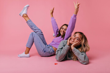 Excited girls in casual spring outfit posing on the floor. Positive best friends fooling around on pastel background.