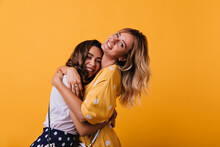 Romantic White Lady In Yellow Dress Embracing Her Sister. Indoor Photo Of Beautiful Trendy Girl Chilling With Best Friend At Weekend Photoshoot.