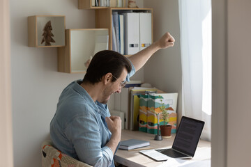 Wall Mural - Overjoyed young man sit at desk at home look at laptop screen celebrate online win or victory, excited male triumph feel euphoric get good news or promotion letter on computer, success concept