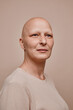 Vertical warm-toned portrait of confident bald woman looking away hopefully while posing against minimal beige background in studio, alopecia and cancer awareness