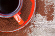 Kawa w czerwonej filiżance ze spodkiem, stojąca na drewnianej poszarzałej desce z rozsypaną, zmieloną kawą. Ujęcie z góry, fragment.