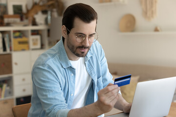 Wall Mural - Young Caucasian man in glasses make online payment or purchase on laptop with credit card from home, millennial male shopping on internet or paying bills using web banking service system on computer