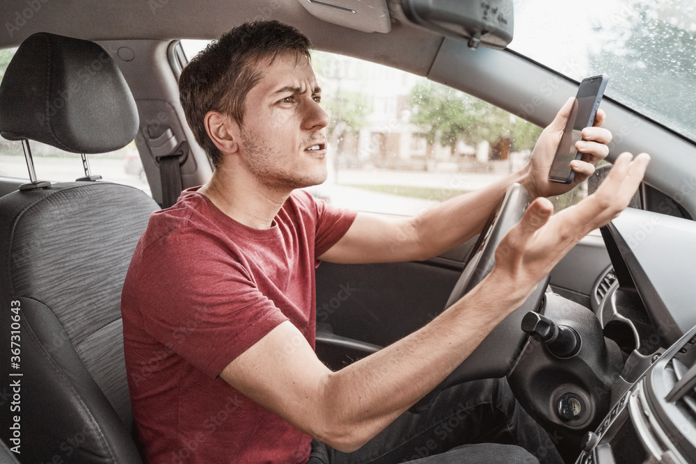 Man behind the wheel of his car loses concentration on the road situation and gets stuck in a smartphone typing SMS in a chat. Risk of a dangerous road accident - obrazy, fototapety, plakaty 