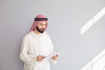 Wall Mural - Attractive smiling arab man writes in clipboard on gray background