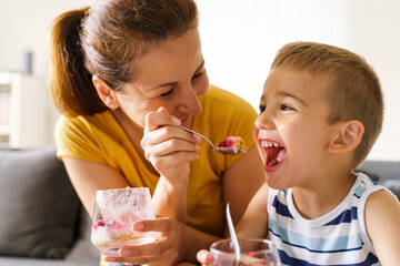 Mother and soon feeding at home in day - Caucasian woman sitting on sofa feed small boy with desert - front view close up - domestic life joy and fun real people bonding childhood concept