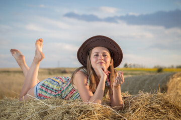 Fototapete - Young girl in a cowboy hat on hay bale. Girl farmer in denim shorts and plaid shirt, in a wheat field in sunset. Happy Woman cowgirl on a farm. Freedom lifestyle.