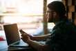 © mykolastock - Closeup portrait of stylish bearded hipster male in hat and eyeglasses sitting in cafe, working on laptop and listening to music