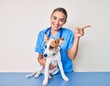 Young beautiful blonde veterinarian woman checking dog health smiling happy pointing with hand and finger to the side