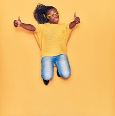 Young beautiful african american woman wearing casual clothes and glasses smiling happy. Jumping with smile on face doing ok sign with thumbs up over isolated yellow background