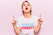 Young Beautiful Blonde Woman Wearing T Shirt With Diversity Word Message Amazed And Surprised Looking Up And Pointing With Fingers And Raised Arms.