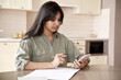 © insta_photos - Indian teen girl holding mobile phone using remote online education app at home. Female student using smartphone online learning in application program writing notes sit at table at home office.