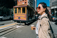 Closeup Side View Portrait Of Smiling Young Beautiful Asian Japanese Woman Walking And Holding Drink Wearing Headphones On Street In City. Cable Car Transportation In San Francisco Commute Lifestyle