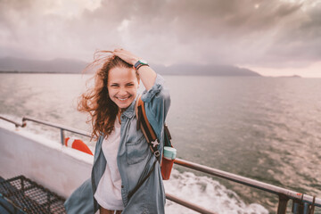 Wall Mural - Happy curly young woman traveler on a ship at the sea against the backdrop of a beautiful landscape. Windy weather and flying hair, adventure travel concept