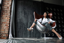 Bottom View Of A Beautiful Woman With Long Hair Sitting In An Aviator Chair In A Loft And With Her Legs Thrown Over The Arm Of A Chair In A Loft