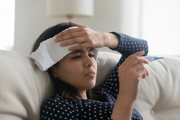 Sick millennial Asian girl sit on couch at home with towel on head measure temperate on thermometer, unhealthy ill young Vietnamese woman suffer from flu or grippe, catch cold, health problem concept