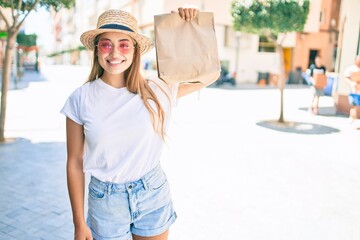 Young beautiful blonde caucasian woman smiling happy outdoors on a sunny day holding delivery paper bag