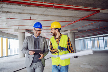 Smiling architect in suit with helmet on head talking to contractor while standing in building in construction process. Architect holding tablet.