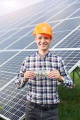 Wall Mural - Portrait of happy man with hundred dollar bill in hand shows thumb a sign like he likes in the background of a solar station. Business and generation concept. Green ecological power energy generation.