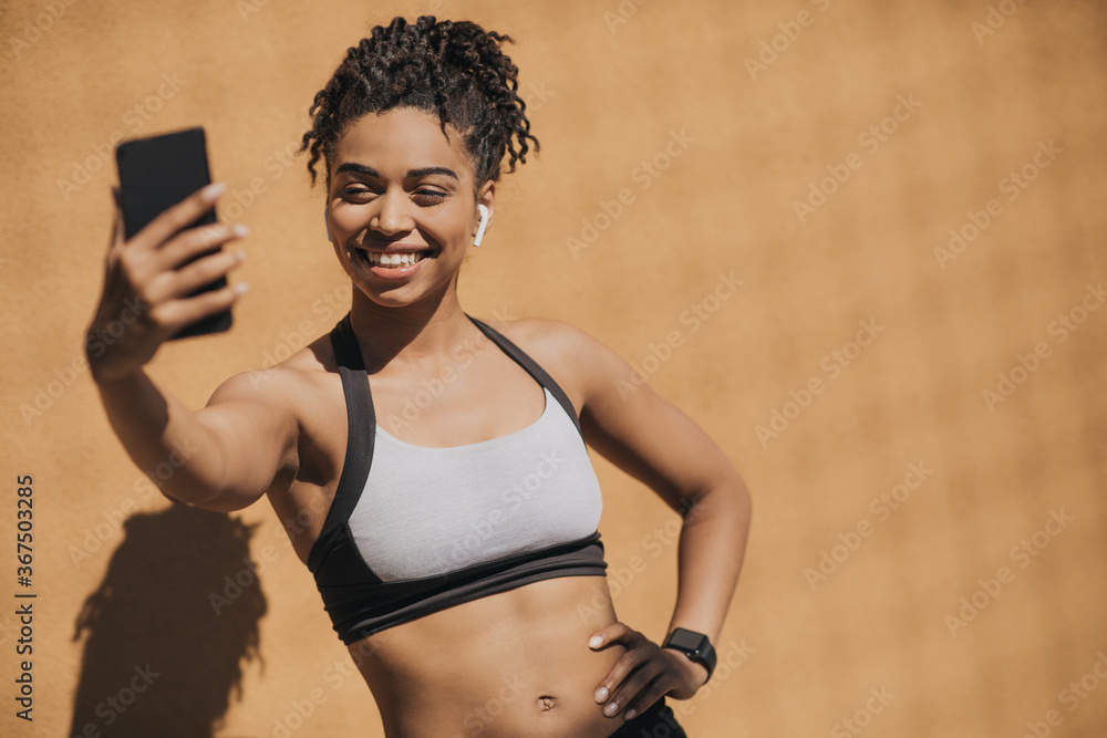 African american young woman in sports bra with wireless headphones and fitness tracker making selfie - obrazy, fototapety, plakaty 