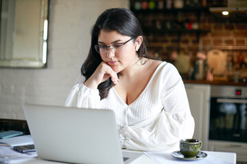 Beautiful serious young woman sales manager with black loose hair and chubby cheeks working remotely, listening to webinar on marketing online using wireless earphones and laptop, wearing glasses