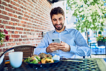 Young businessman using smartphone while eating breakfast outdoor in cafe