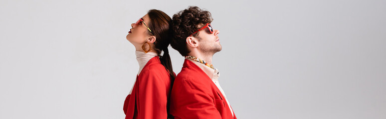 Wall Mural - panoramic shot of trendy couple in red blazers and sunglasses standing back to back isolated on grey