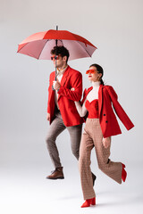 Wall Mural - full length view of trendy couple in red blazers and sunglasses running with umbrella on grey