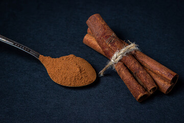 Cinnamon on a black background. A spoonful of ground cinnamon lies next to two cinnamon sticks tied with jute string.