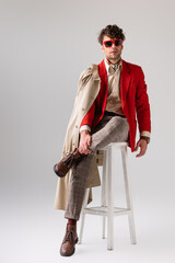 Wall Mural - handsome, confident man in stylish autumn clothes touching leg while sitting on stool on grey