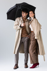 Wall Mural - full length view of young couple in fashionable autumn clothes posing under umbrella on grey