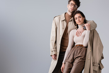 Wall Mural - trendy man in trench coat hugging fashionable woman while looking at camera isolated on grey