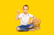 © Olga Mandrik - The schoolboy sits on a yellow background next to a mountain of books and points his finger up. Caucasian boy in a white T-shirt and ripped jeans. Concept of education, back to school. Knowledge Day