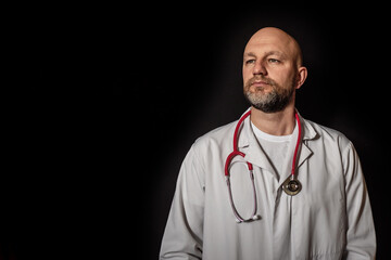 Portrait of a bald doctor with beard in white uniform and red stethoscope around his neck, dark background. Man looking away from camera.