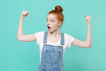 Wall Mural - Shocked young readhead girl in casual denim clothes posing isolated on blue turquoise background studio portrait. People sincere emotions lifestyle concept. Mock up copy space. Showing biceps muscles.