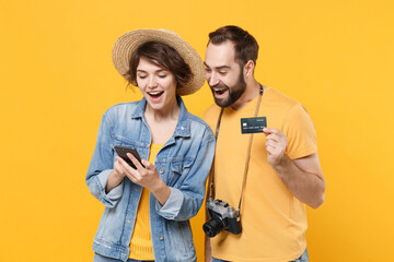 Wall Mural - Excited young tourists couple friends guy girl in summer clothes isolated on yellow background. Passenger traveling abroad on weekends. Air flight journey concept. Hold mobile phone, credit bank card.