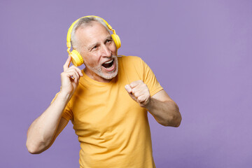 Cheerful elderly gray-haired mustache bearded man in casual yellow t-shirt posing isolated on violet wall background studio portrait. People lifestyle concept. Listen music with headphones, sing song.