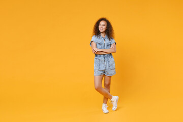 Wall Mural - Full length portrait of smiling young african american girl in denim clothes isolated on yellow background studio. People lifestyle concept. Mock up copy space. Holding hands crossed looking aside.