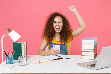 Happy african american girl employee in office sit isolated on pink background. Achievement business career. Education in school university college concept. Using mobile phone, doing winner gesture.