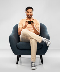 people, technology and furniture concept - happy smiling young indian man playing game on smartphone sitting in chair over grey background