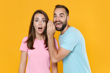 Shocked couple friends guy girl in blue pink t-shirts isolated on yellow background studio portrait. People lifestyle concept. Mock up copy space. Whispers gossip and tells secret with hand gesture.